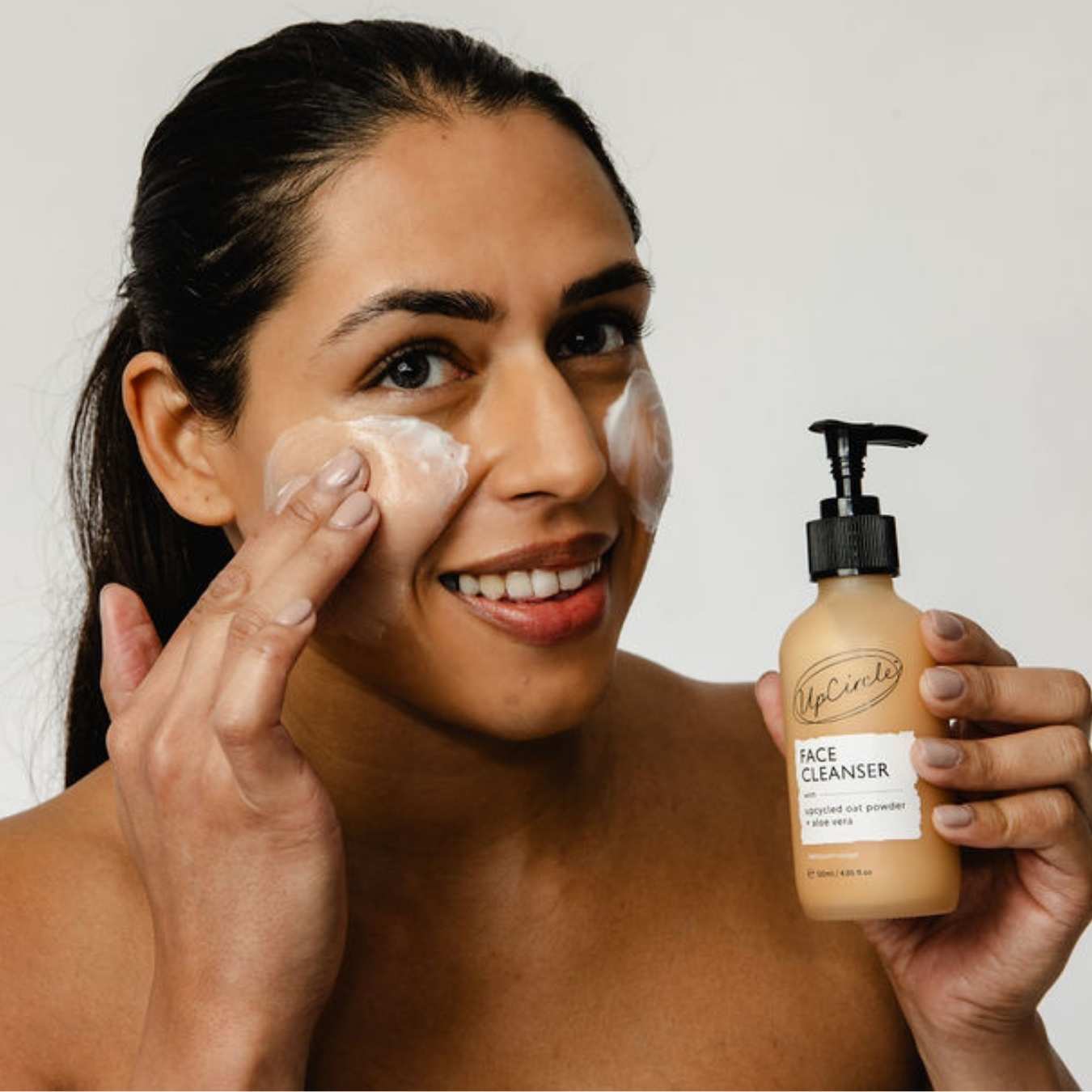 Woman applying facial cleanser to her face with a bottle of the product in her other hand.